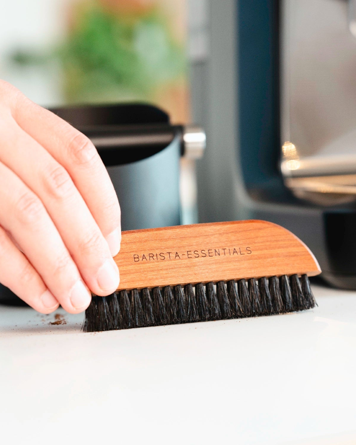 Hand, die mit einer Holzbürste Kaffeesatz von einer Arbeitsplatte wischt.